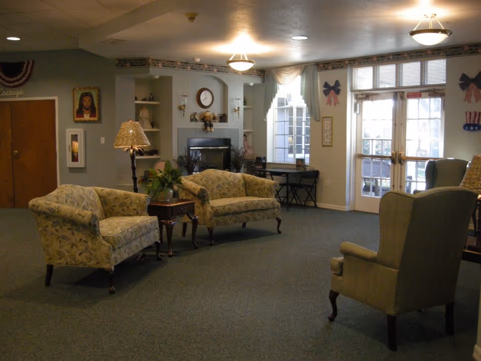 A cozy senior living facility common area with floral patterned sofas, an armchair, a wooden side table with a plant, a floor lamp, a fireplace with a clock above it, and large windows with curtains letting in natural light. The room has carpeted flooring and decorative wall hangings including patriotic bows and a portrait.