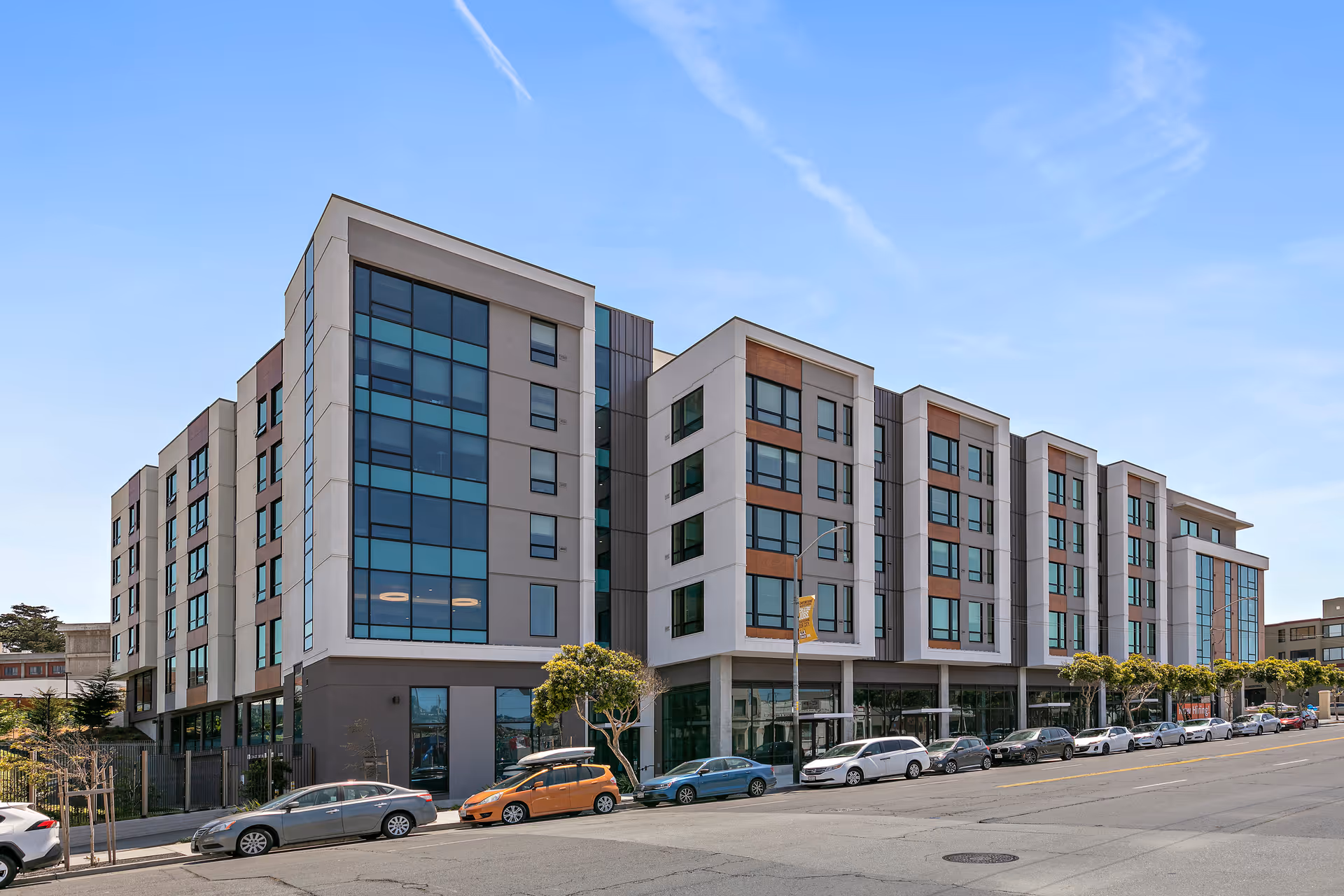 Exterior view of a modern multi-story assisted living and memory care facility with large windows, several parked cars along the street, and a clear blue sky.
