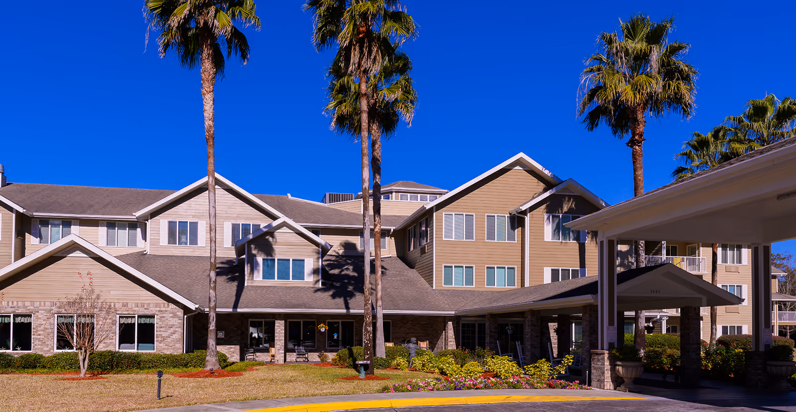 Exterior view of a multi-story senior living facility building with beige siding and brick accents, surrounded by palm trees and landscaped gardens under a clear blue sky.
