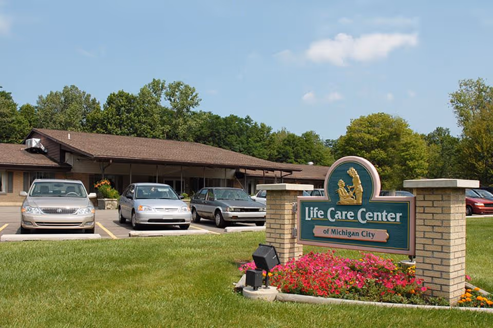 Front sign for Life Care Center of Michigan City with a flowerbed, parked cars, and the facility building in the background.