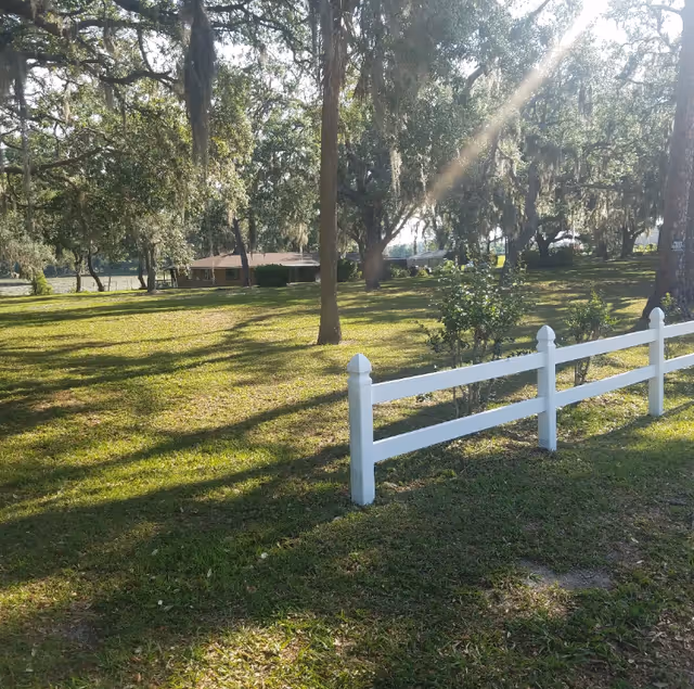 A sunny outdoor scene with a grassy area, several large trees casting shadows, a white wooden fence in the foreground, and a building partially visible in the background.