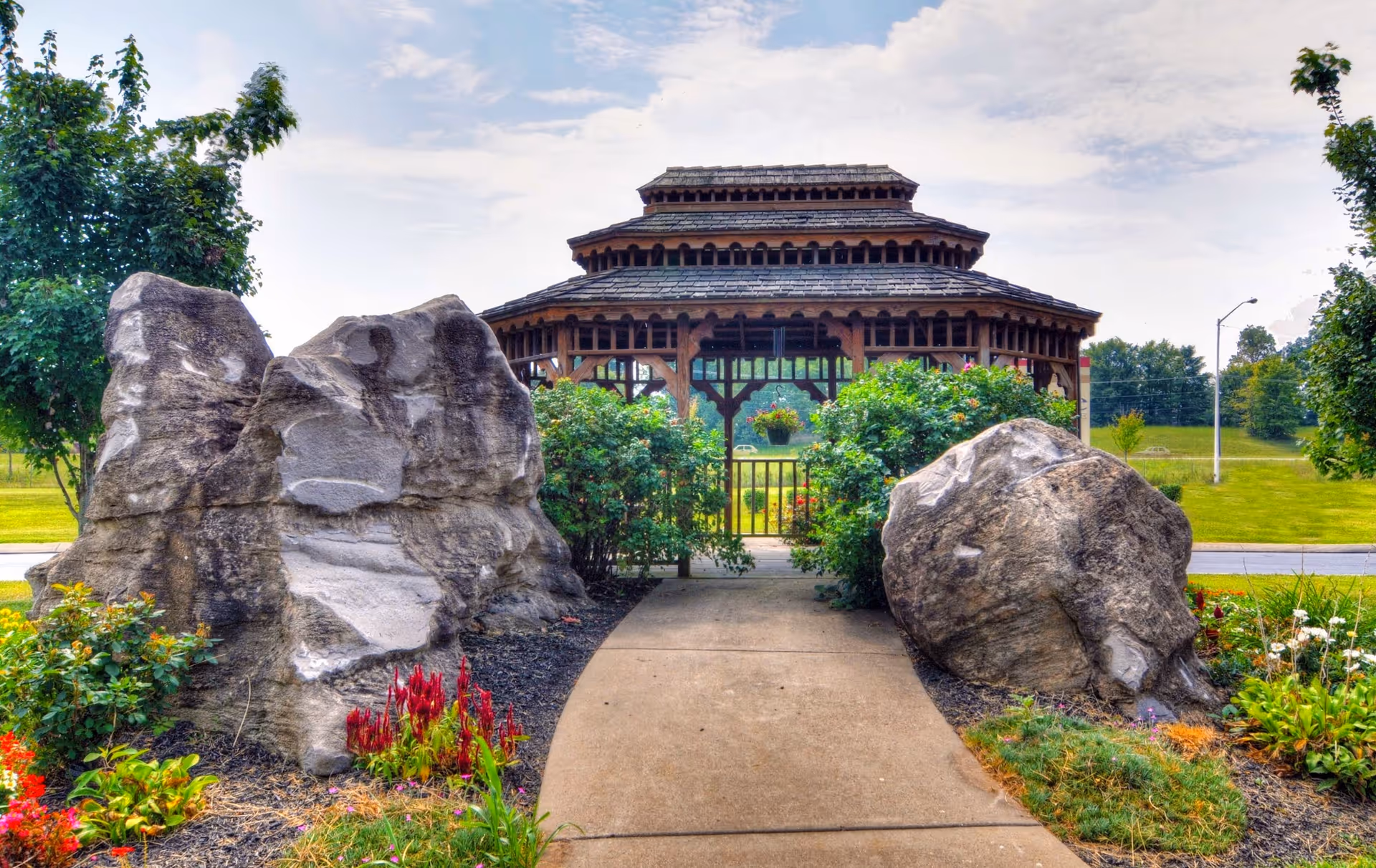 A paved path leads between large decorative rocks and flower beds to a wooden gazebo surrounded by greenery.