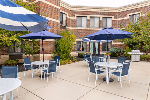 Outdoor patio area at Elderwood Assisted Living at Cheektowaga with several round white tables and blue chairs. Each table has a blue umbrella providing shade. The patio is surrounded by a brick building with windows and some greenery including bushes and small trees.