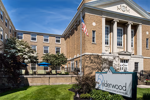 Front exterior of the Elderwood Assisted Living at Hamburg building with a lawn, American flag, and facility sign.