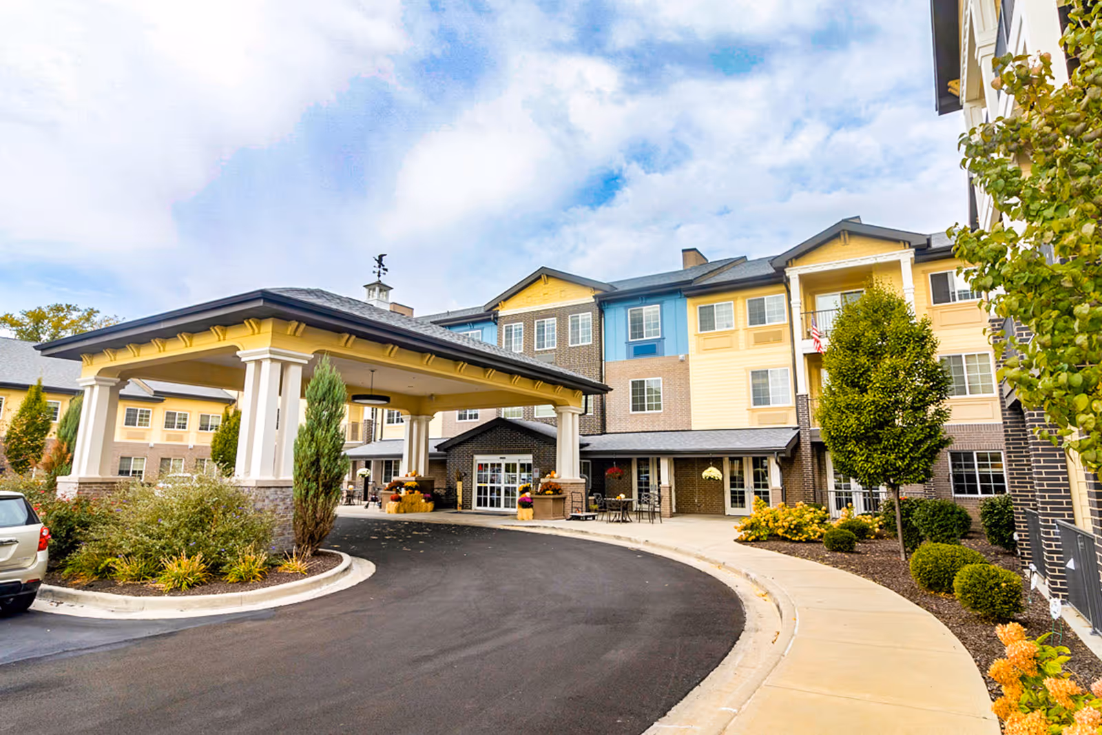 Exterior view of Independence Village of Carmel showing a multi-story building with a covered entrance driveway, landscaped bushes, trees, and a clear sky with some clouds.