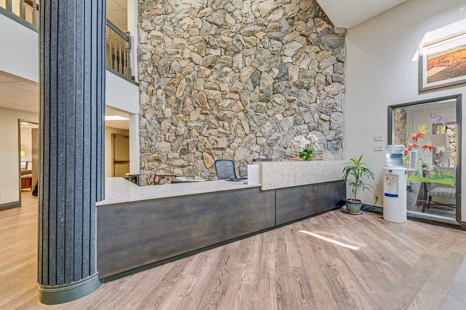 Reception area with a large stone accent wall behind a modern reception desk. The desk has a marble countertop and dark paneling. There is a tall column to the left, a water cooler and potted plants to the right, and a framed picture on the wall above the water cooler. The floor is wood with a light finish.