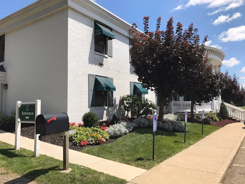 Front exterior of a white two-story building with green window awnings, landscaped flowerbeds, a mailbox, and an entrance sign.