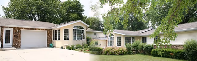 Exterior view of a single-story residential building with a garage on the left side, multiple windows, and surrounded by green trees and shrubs under a clear sky.