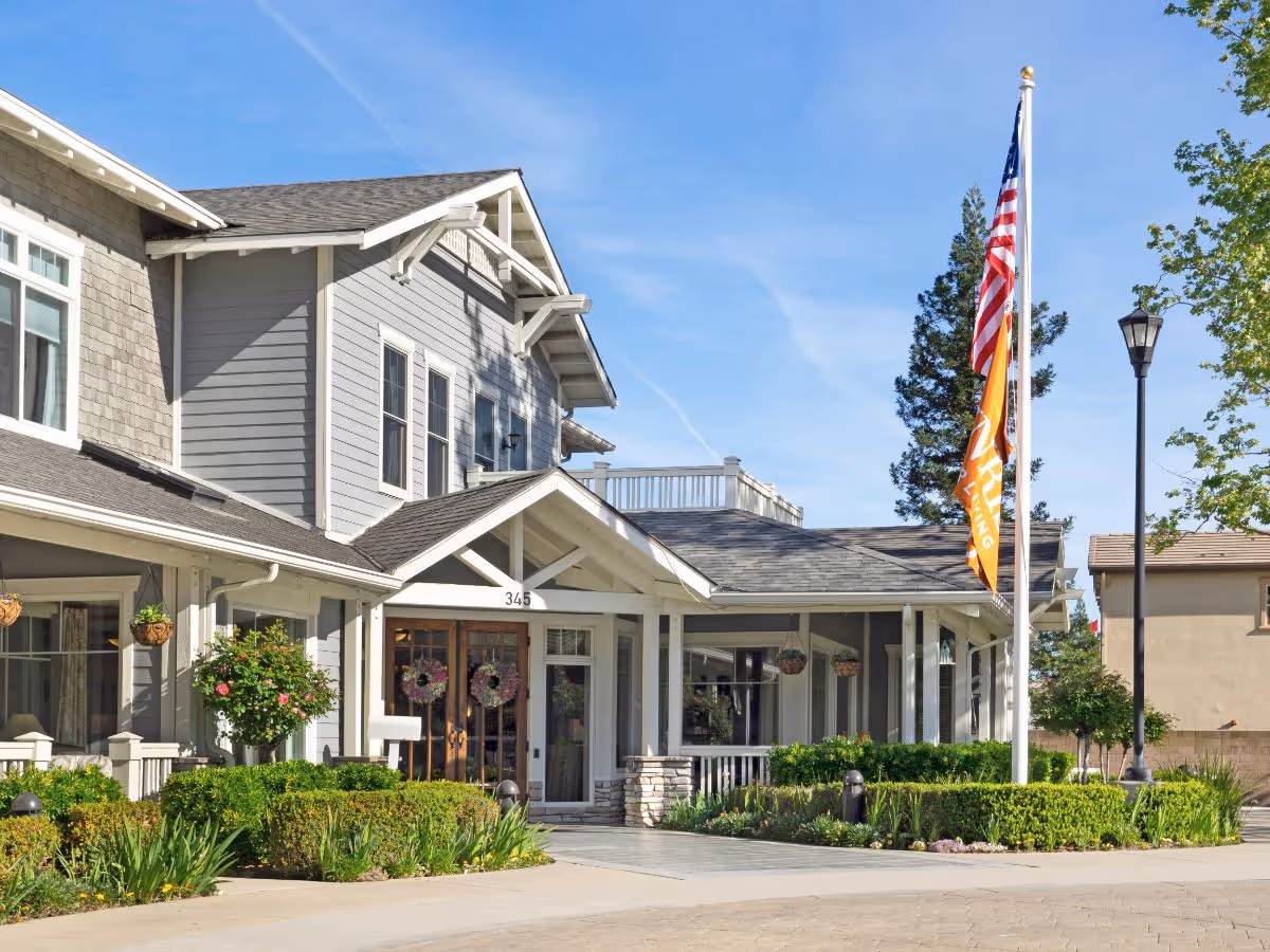 Exterior view of a senior living facility building with gray siding, white trim, and a covered entrance. There are two wreaths on the glass double doors, well-maintained landscaping with bushes and flowers, an American flag and an orange flag on a flagpole, and a street lamp nearby under a clear blue sky.
