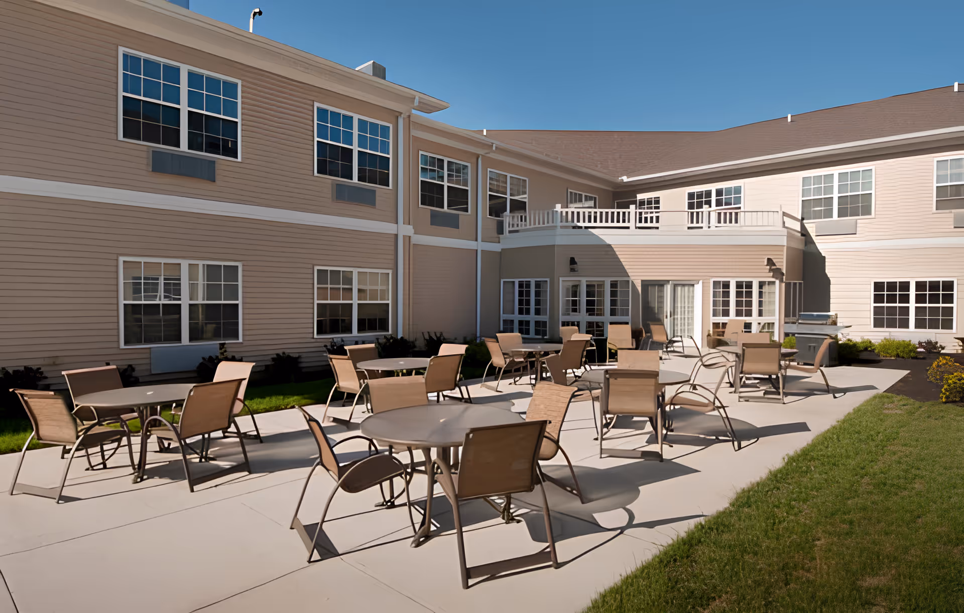 Outdoor patio area at Heathwood Assisted Living and Memory Care with multiple round tables and chairs arranged on a concrete surface, surrounded by a two-story beige building with many windows under a clear blue sky.