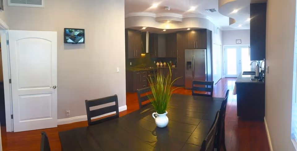 Interior view of a modern kitchen and dining area with dark wood cabinets, stainless steel refrigerator, and a black dining table with chairs. A white vase with green plants is placed on the table. The room has wooden flooring and light-colored walls, with a door and windows allowing natural light.