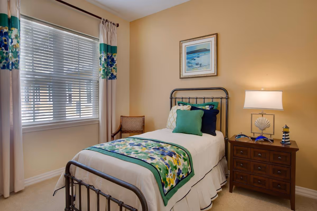 A cozy bedroom with a single metal bed frame, white bedding, and a colorful floral throw blanket and pillows. There is a wooden nightstand with a lamp, decorative items including a shell and lighthouse figurine, and a framed picture of a coastal scene on the beige wall. A window with white blinds and floral curtains is on the left side, and a wooden chair is placed in the corner.