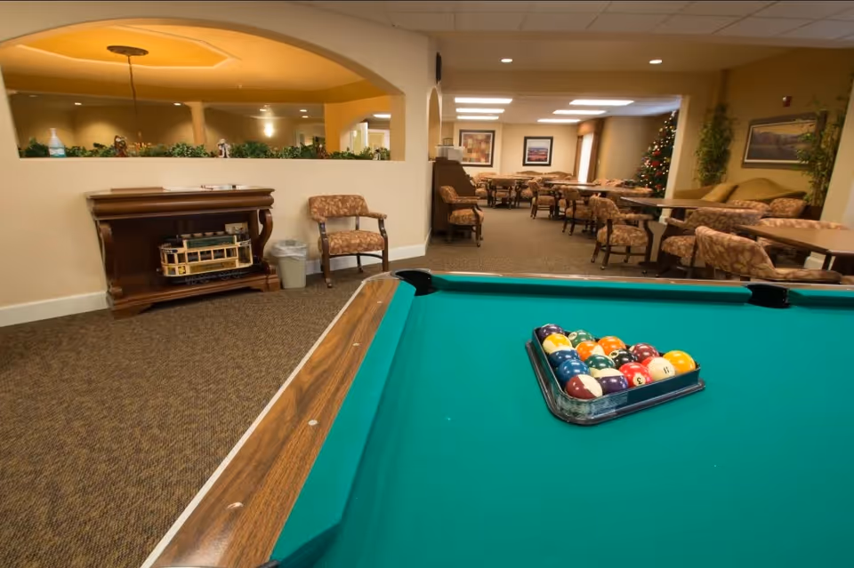 A billiards table with racked pool balls in the foreground and a seating/dining common area with tables and chairs in the background.