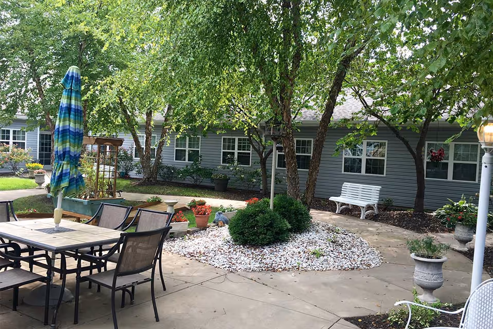Outdoor patio area with a table and chairs, a closed blue and green striped umbrella, trees, potted plants, a white bench, and a building with multiple windows in the background.