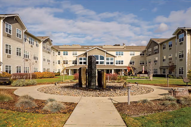 Outdoor courtyard area of a senior living facility with a circular stone fountain in the center, surrounded by a paved walkway, landscaped grass, and shrubs. The three-story beige building with multiple windows and a covered entrance is visible in the background under a partly cloudy sky.