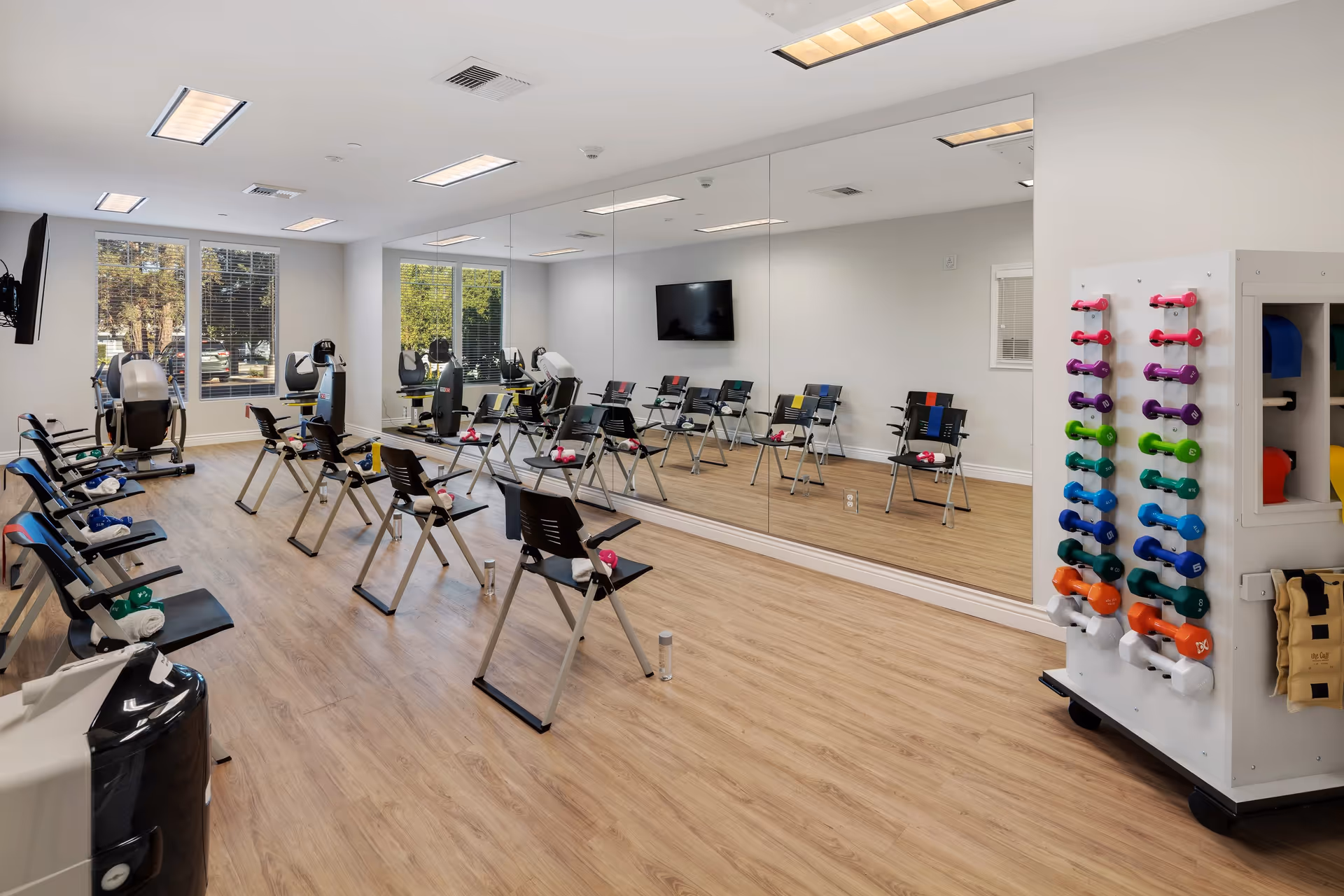 A bright fitness room with wooden flooring, several black chairs arranged in rows, each with a small towel and a dumbbell on the seat. A large wall mirror reflects the room, and a flat-screen TV is mounted on the wall. A rack with colorful dumbbells is visible on the right side, and large windows let in natural light with a view of trees outside.