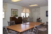 A room with a rectangular wooden table surrounded by metal chairs with cushioned seats. The room has white walls, two windows with curtains, a piano against one wall with a framed picture above it, and a door in the background. The ceiling has fluorescent lighting.