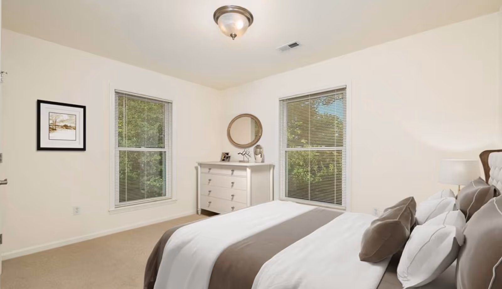 A bright bedroom with a neatly made bed featuring white and brown bedding, a white dresser with a round mirror above it, two windows with blinds showing greenery outside, a framed picture on the wall, and a ceiling light fixture.