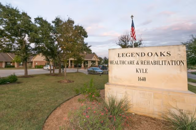 Outdoor view of Legend Oaks Healthcare & Rehabilitation Kyle facility sign with an American flag on top, surrounded by grass, plants, and trees, with the building and parked cars visible in the background under a partly cloudy sky.