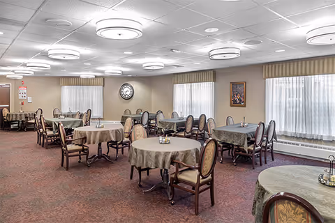 A spacious dining room with several round tables covered with tablecloths and surrounded by upholstered chairs. The room has large windows with sheer curtains and valances, a carpeted floor, and ceiling lights evenly spaced across the ceiling. There is a clock on the wall and a framed picture hanging beside the windows.