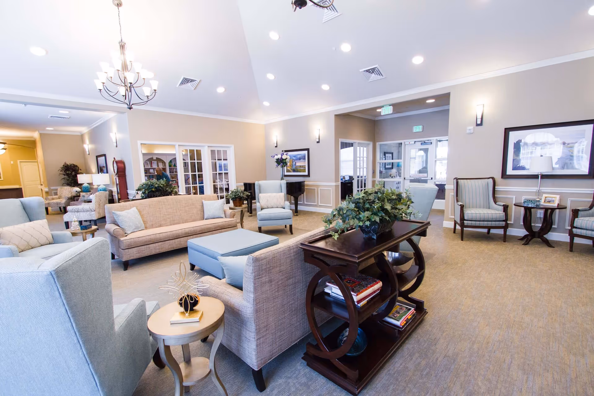 A spacious and well-lit living room area in a senior living facility featuring beige and light blue upholstered sofas and armchairs arranged around a central ottoman. There is a dark wooden console table with plants and books, wall art, and a chandelier hanging from the ceiling. The room has beige walls with white trim and multiple exit signs above doors.