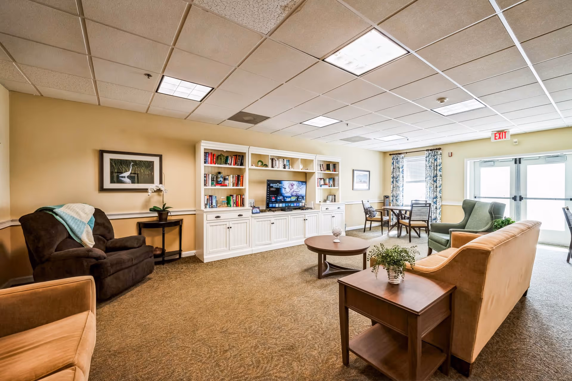 A cozy senior living common area with beige walls and carpeted floor. The room features a brown armchair with a light blue and white blanket draped over it, a tan sofa, a green armchair, and a wooden coffee table with a small plant on it. There is a white built-in bookshelf and entertainment center with a TV displaying a streaming service. A small dining table with four chairs is near the window, which is adorned with floral curtains. The room is well-lit with ceiling lights and natural light coming through glass doors marked with an exit sign.