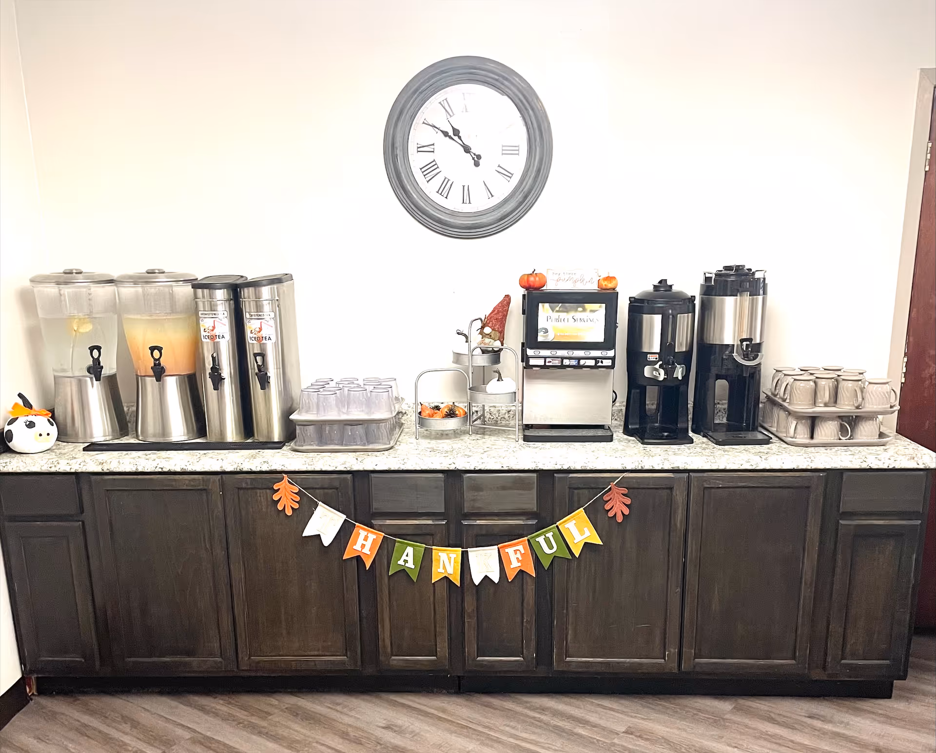 A beverage station with multiple drink dispensers including iced tea, coffee, and water on a granite countertop. There are stacks of plastic cups and ceramic mugs. Above the station is a round wall clock showing the time as 11:55. A decorative banner spelling 'THANKFUL' is hanging on the front of the dark wooden cabinets below the countertop. Small seasonal decorations are placed on the counter.