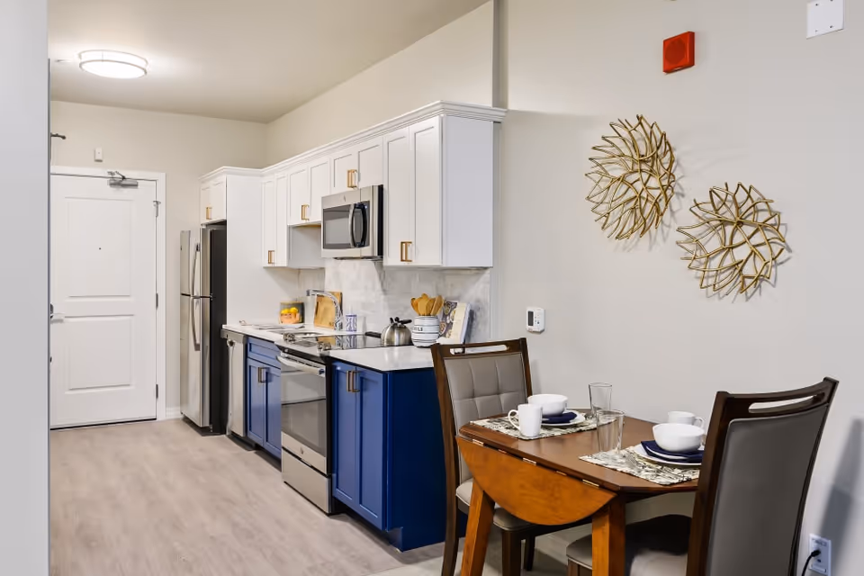 Modern kitchen with white upper cabinets and blue lower cabinets, stainless steel appliances including a refrigerator, oven, and microwave. A small wooden dining table set for two with white dishes and glasses is positioned next to the kitchen. Two decorative gold wall hangings are on the wall above the dining table.