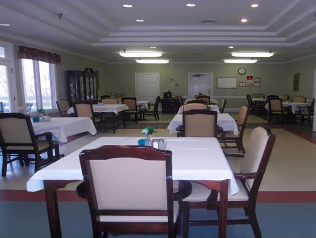 A dining room with multiple tables covered in white tablecloths and surrounded by wooden chairs with beige cushions. The room has large windows on the left side letting in natural light, a cabinet against the far wall, and a clock and bulletin board on the back wall.