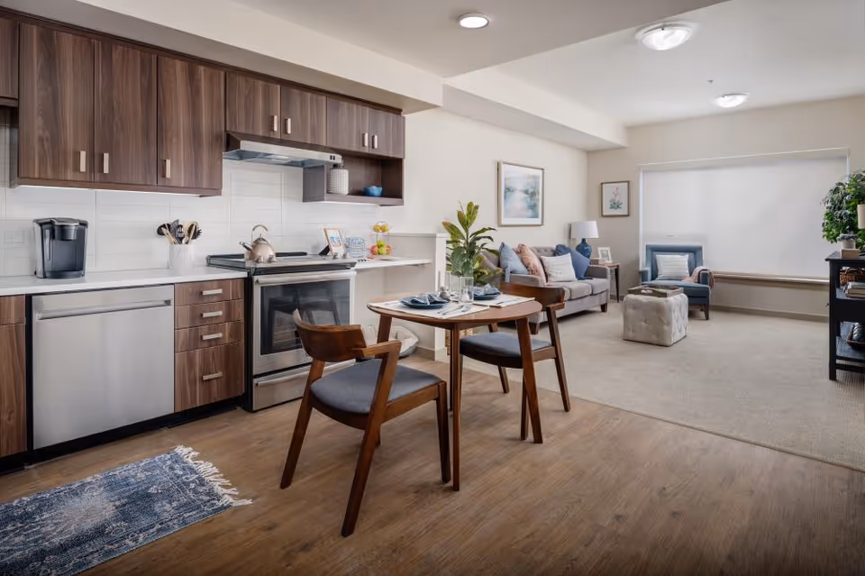 A modern open-concept living space featuring a kitchen with wooden cabinets, stainless steel appliances, and a small dining table set for two. The living room area includes a gray sofa with cushions, a blue armchair, a tufted ottoman, a side table with a lamp, and framed artwork on the walls. The room has wood flooring in the kitchen and dining area, and carpet in the living area, with a large window covered by a white blind.