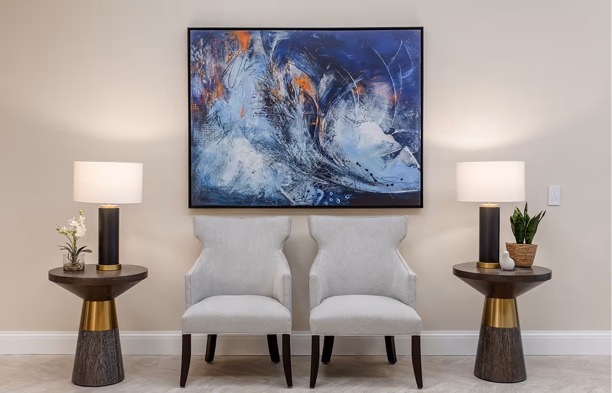 Two light gray upholstered chairs placed side by side against a beige wall, with a large abstract painting featuring blue, white, and orange tones hanging above them. On each side of the chairs is a round wooden side table with a black and gold base, each holding a modern table lamp with a white shade. The left table also has a small white orchid plant, and the right table has a potted green plant and a small decorative white object.