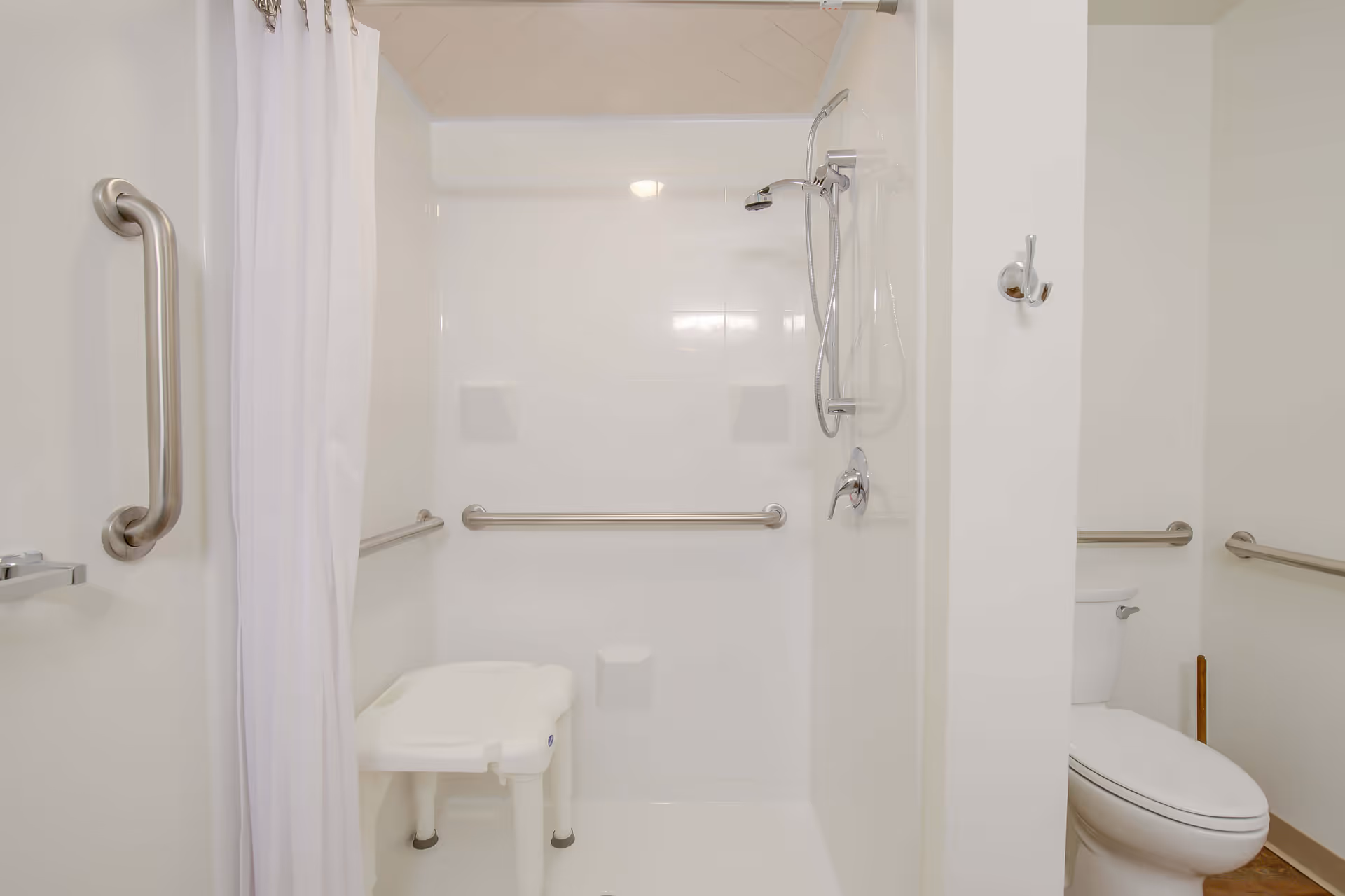 A clean, accessible bathroom featuring a walk-in shower with a white shower chair and multiple stainless steel grab bars. There is a white shower curtain partially drawn to the left, and a toilet with grab bars on the right side of the image.