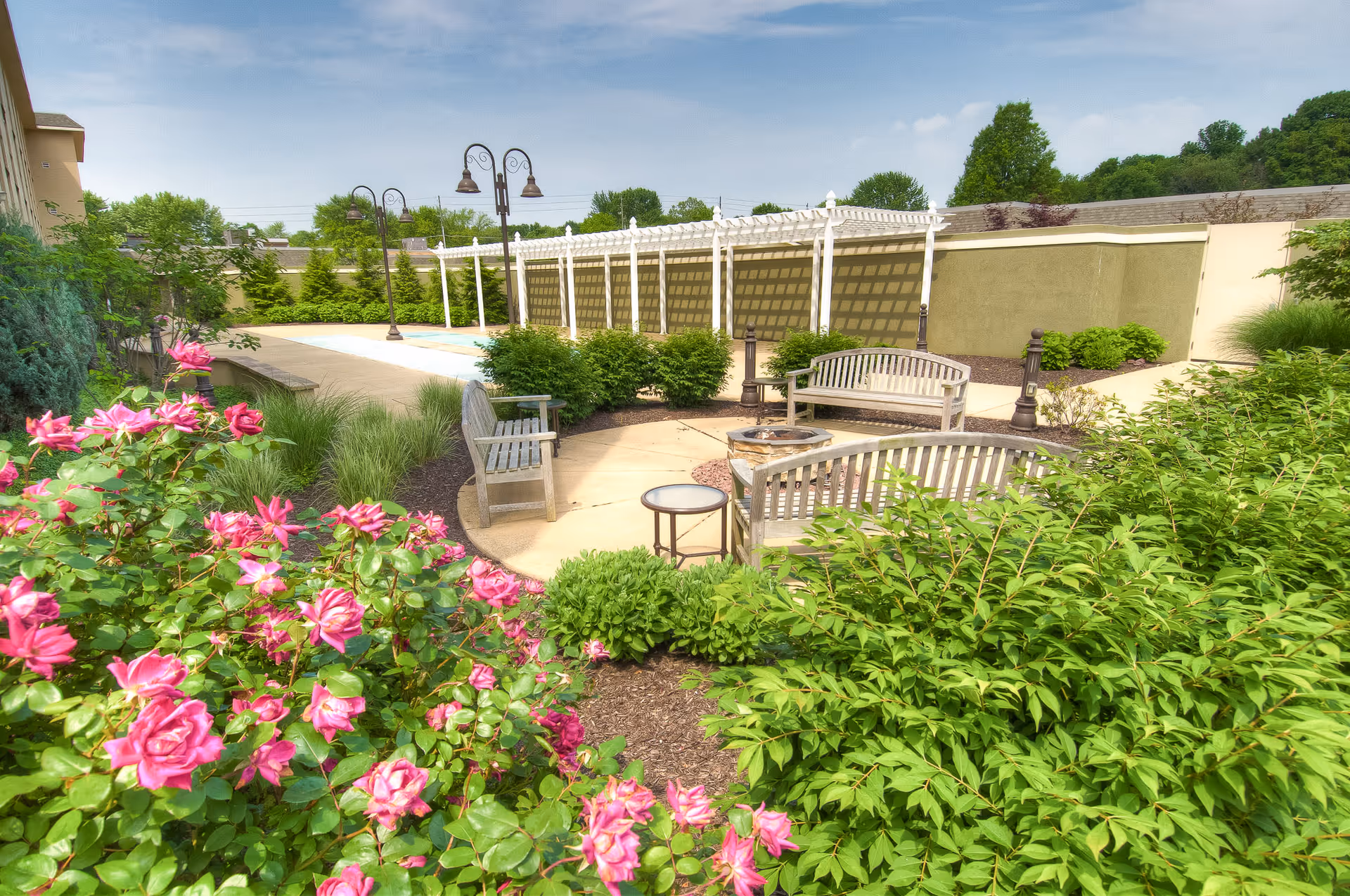 Outdoor garden area at Westside Garden Plaza with blooming pink roses in the foreground, green shrubs, wooden benches arranged around a fire pit, a small round glass table, and a white pergola structure along a paved walkway under a clear blue sky.