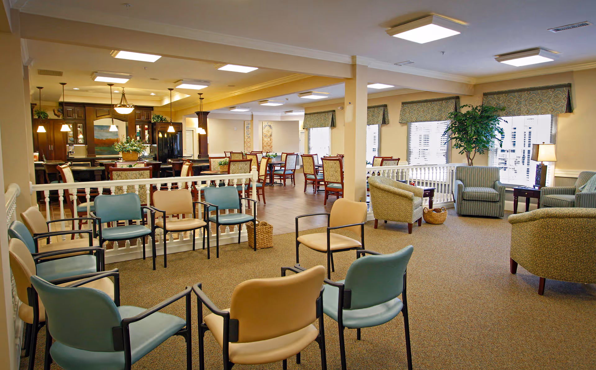 A spacious senior living common area with a circle of chairs in the foreground and a dining area with tables and chairs in the background. The room is well-lit with ceiling lights and natural light from windows with valances. There are also comfortable armchairs, side tables, a large indoor plant, and a kitchen area visible in the back.