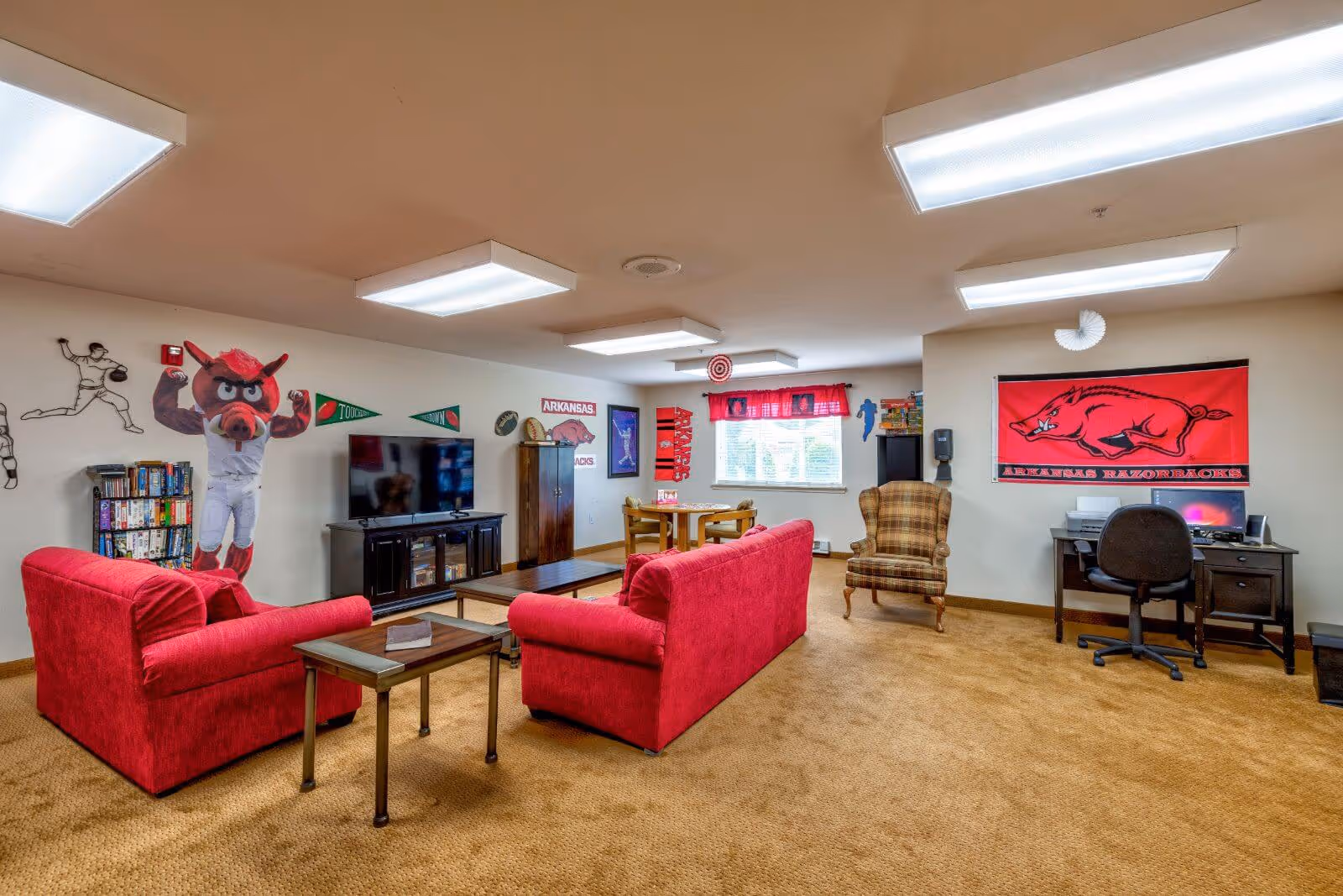 Communal lounge with red sofas facing a TV, game shelves, a desk and Arkansas Razorbacks decor on the walls.