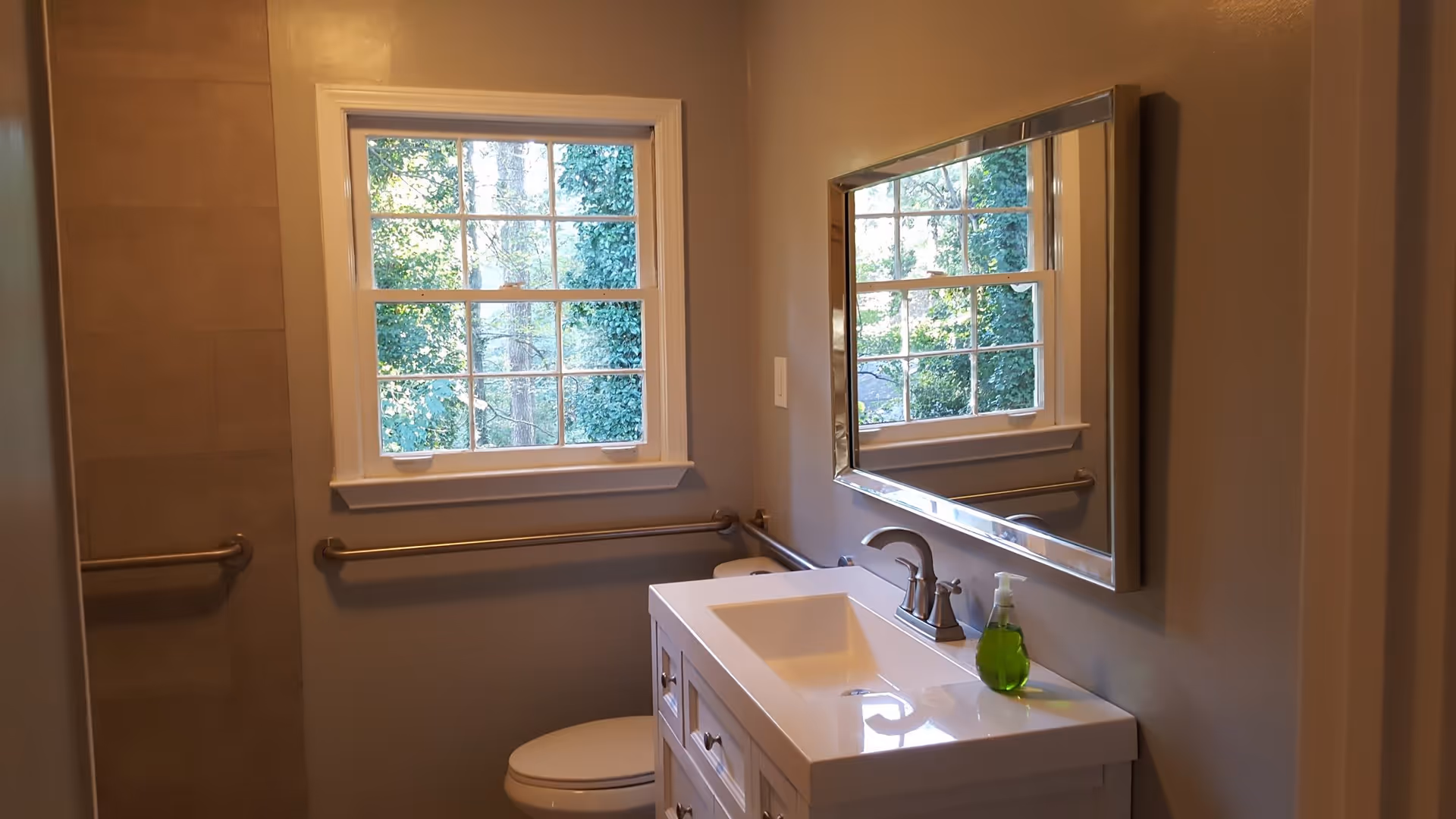 A small bathroom with a white vanity and sink topped with a green liquid soap dispenser. Above the sink is a large rectangular mirror reflecting a window with a view of green trees outside. The bathroom has beige walls and a toilet partially visible next to the vanity. There are metal grab bars installed on the walls near the toilet and window.