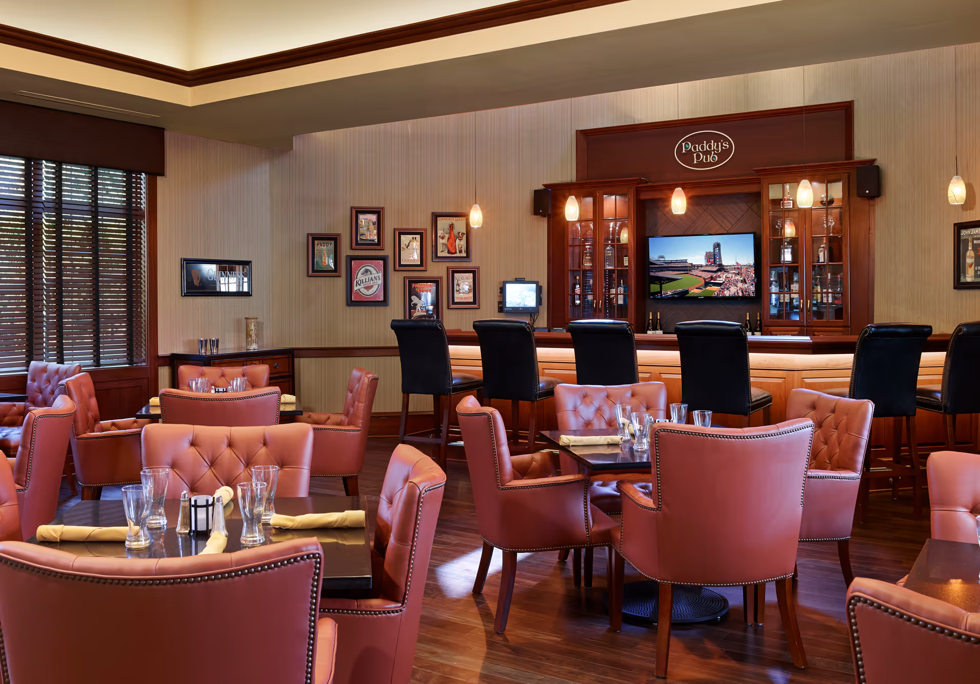 Interior view of a cozy pub-style dining area with several tables and pink cushioned chairs. A wooden bar with high black chairs is visible in the background, with a sign above reading 'Paddy's Pub'. The walls are decorated with framed pictures and a television screen is mounted behind the bar. Warm lighting and wooden flooring create a welcoming atmosphere.
