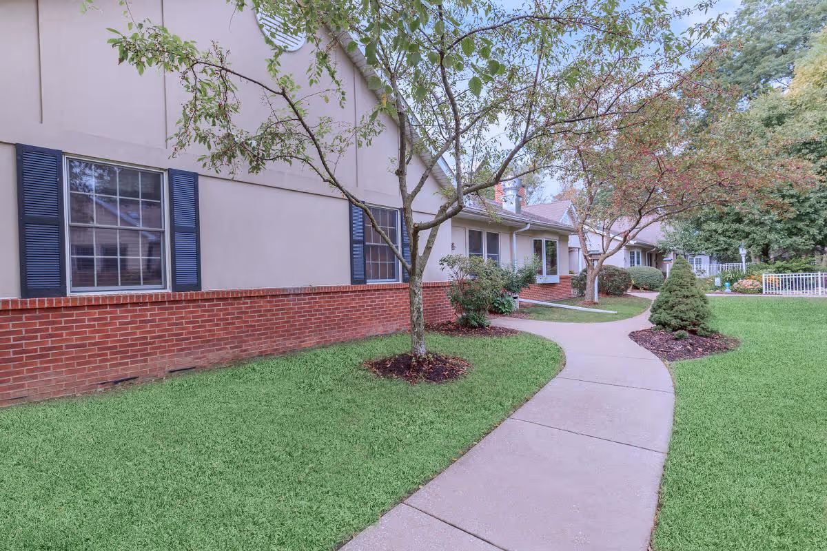 Curved concrete pathway leading alongside a building with beige walls and red brick lower exterior. The path is bordered by green grass, small trees, and shrubs. The building has multiple windows with dark shutters, and there are more trees and greenery in the background.