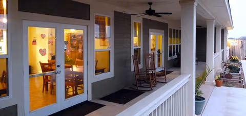 Covered exterior porch with rocking chairs and lit windows showing a dining area through French doors.