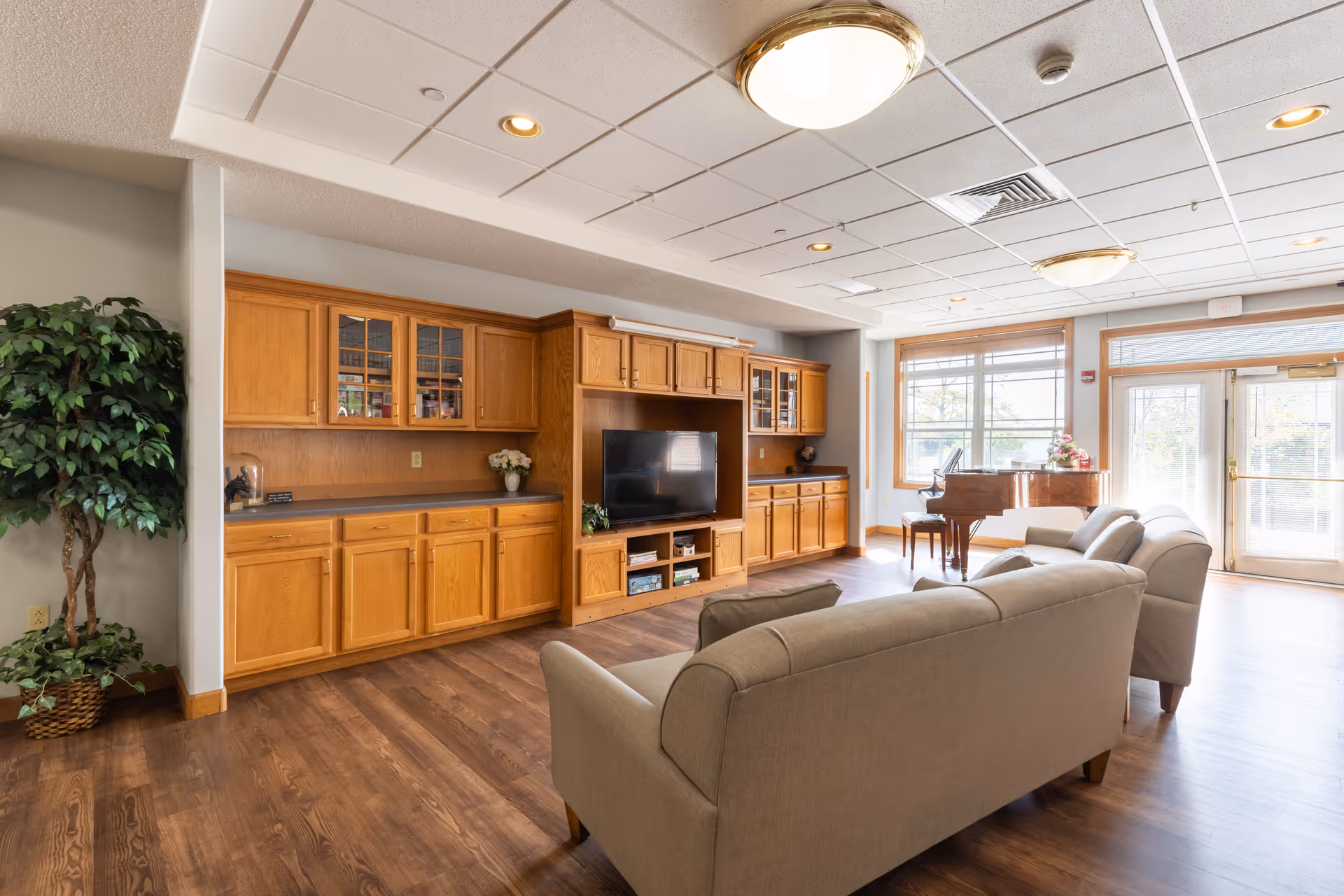 A bright and spacious living room area with wooden flooring, beige sofas, a large wooden entertainment center with a flat-screen TV, and a grand piano near large windows letting in natural light. There is also a potted plant in the corner and ceiling lights.