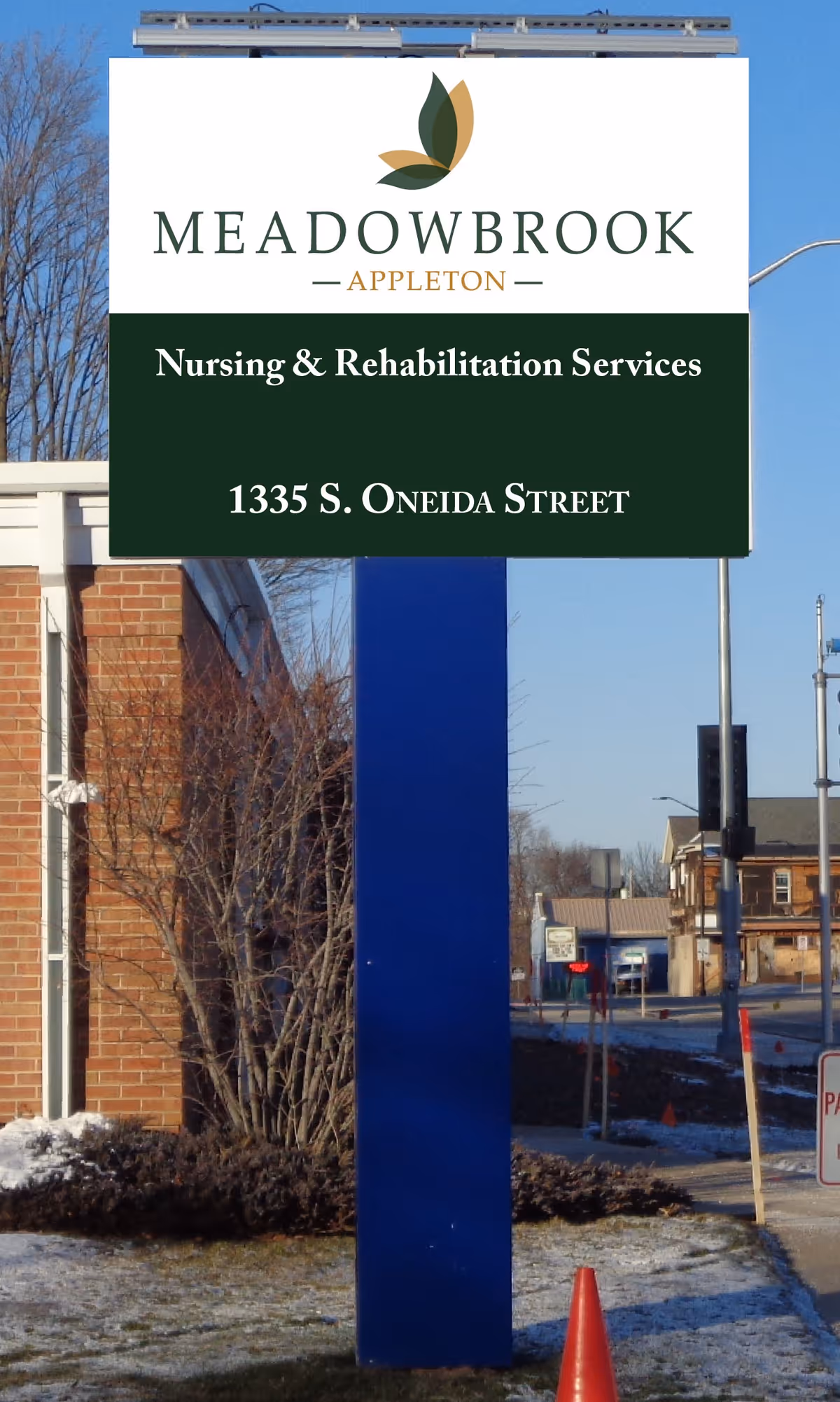 Outdoor sign for Meadowbrook Appleton Nursing & Rehabilitation Services located at 1335 S. Oneida Street, mounted on a blue post near a brick building with some snow on the ground and leafless trees in the background.