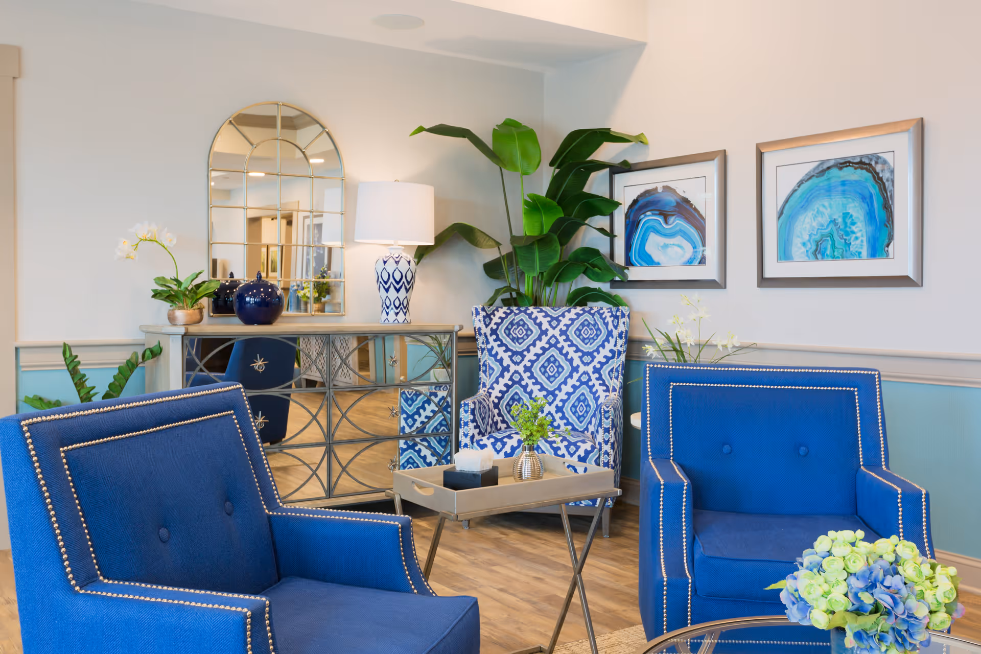 A cozy seating area in a senior living facility featuring two blue armchairs with nailhead trim, a patterned blue and white accent chair, a small wooden tray table with decorative items, a large green plant, a table lamp with a blue and white patterned base, a mirrored console table, and two framed blue abstract art pieces on the wall.