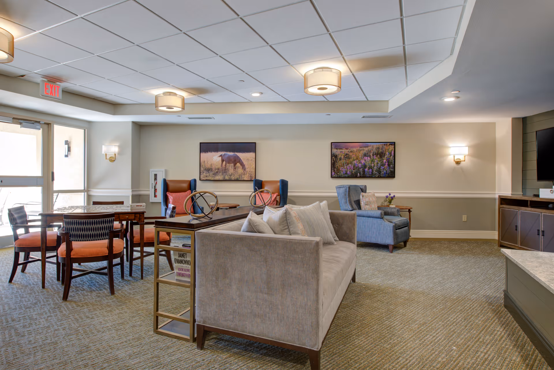 A cozy senior living facility common area with a beige sofa, blue armchairs, and a wooden table with chairs near a glass door. The walls are decorated with framed pictures of a horse and wildflowers. The room is well-lit with ceiling lights and wall sconces, and there is a television mounted on the right wall.