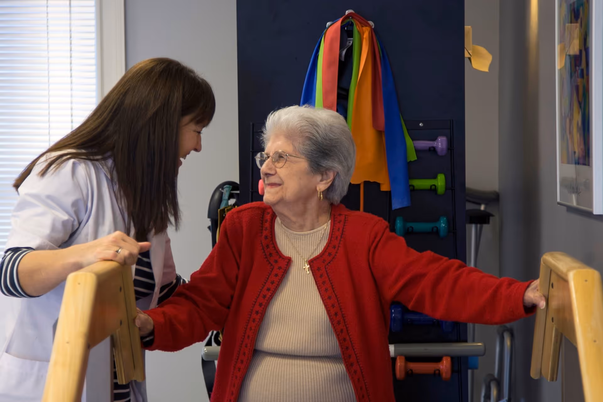 An elderly woman wearing a red cardigan and glasses is holding onto parallel bars for support while a female caregiver or therapist in a white coat smiles and encourages her during a physical therapy or rehabilitation session in a facility.