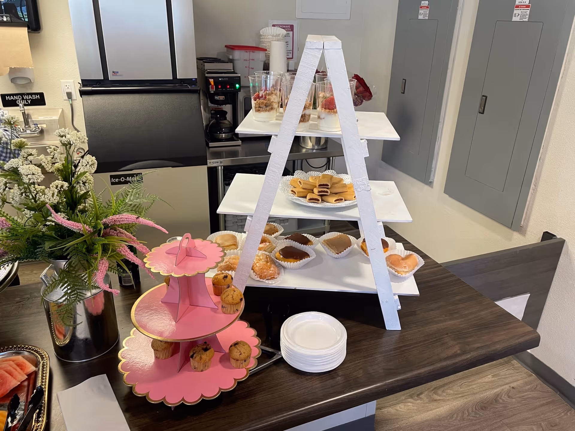 Tabletop display of pastries and desserts on tiered stands in a kitchenette area with flowers, plates, and a coffee machine in the background.