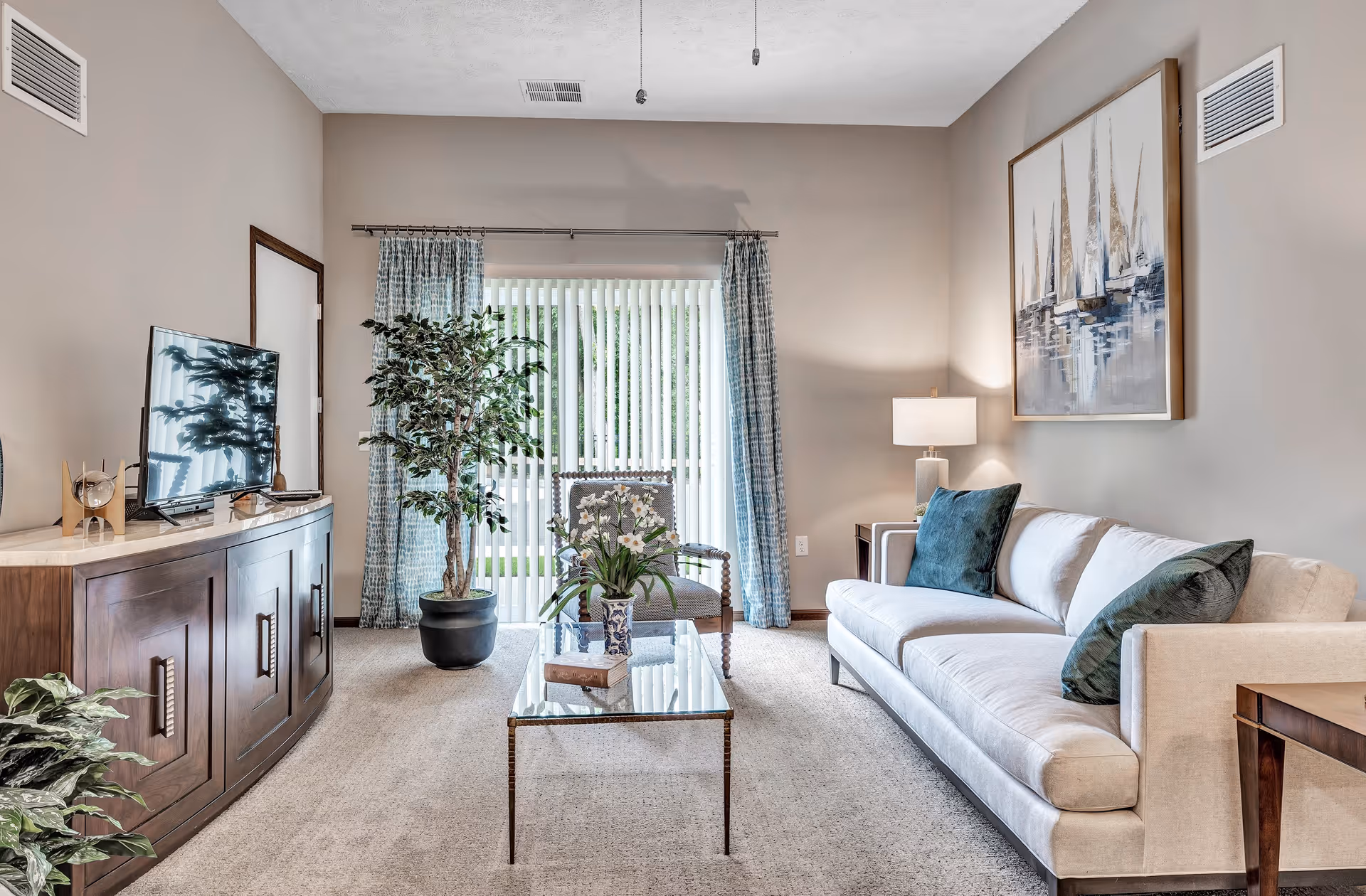 Bright, well-decorated living room with a beige sofa, glass coffee table, TV on a wooden console, potted plants, and sliding vertical-blind doors.