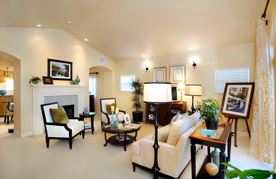 A bright and cozy living room with beige walls and carpet. The room features a white fireplace with a framed painting above it, two white armchairs with dark wood frames, a round wooden coffee table with candles and picture frames, a beige sofa with cushions, a wooden side table with a potted plant and books, and several lamps providing warm lighting. There are two small windows with blinds and a framed picture on an easel near a window with patterned curtains.