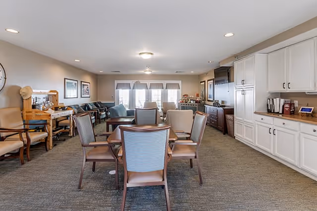 A spacious common area in a senior living facility featuring a central table with six chairs, additional seating along the walls including armchairs and sofas, large windows with blinds allowing natural light, and white cabinetry with a countertop on the right side.