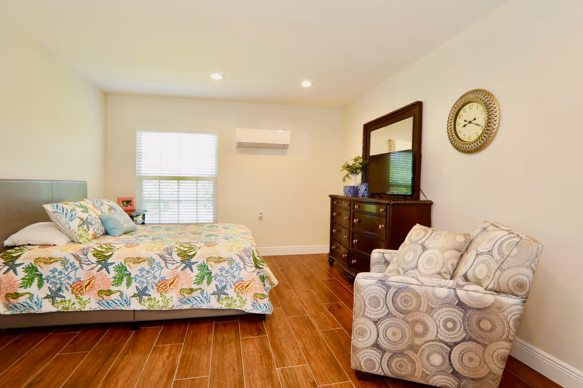 A bright bedroom with a bed covered in a colorful sea-themed quilt, a wooden dresser with a large mirror and a TV on top, a patterned armchair, a wall clock, and a window with blinds letting in natural light.