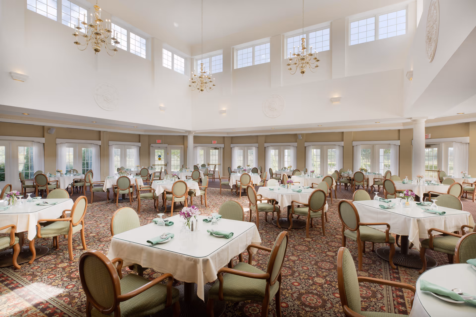 A spacious and elegant dining room with multiple round and square tables covered with white tablecloths, each set with green napkins and small flower arrangements. The room features high ceilings with large windows near the top, allowing natural light to fill the space. There are several chandeliers hanging from the ceiling, and the floor is covered with a patterned carpet. The walls have large windows and doors with beige curtains.
